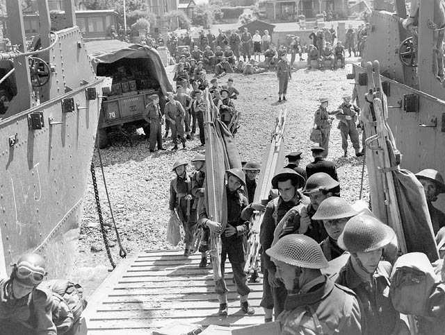 Photographie en noir et blanc – Des hommes en tenue de combat transportant de l’équipement (civières, etc.) avancent vers la rampe d’une péniche de débarquement. On aperçoit derrière d’autres hommes les observant. Il y a un camion militaire.